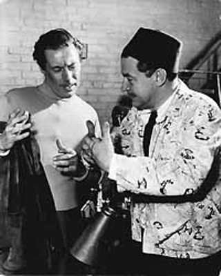 Director Preston Sturges -- who wears a red fez so that the crew can quickly locate the director -- shows Rex Harrison a new technique in thumb-twiddling during the filming of <i>Unfaithfully Yours</i>.