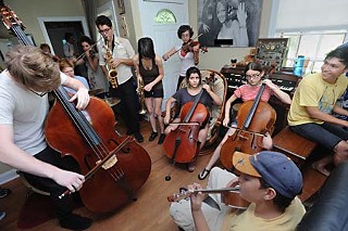 Crammed into the living room and spilling into the hall and kitchen are 14 classical musicians, their instruments, and their cases. The strings are huddled by the front door; percussion and horns stand over the couch.